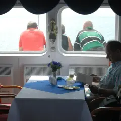 Rejs PKP Gdynia - Hel. Cena biletu? Mężczyzna czyta w restauracji na statku, podczas gdy inni pasażerowie patrzą na morze.