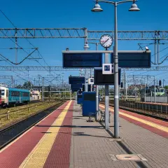 Peron kolejowy z pociągami, tablicami informacyjnymi i zegarem.