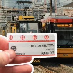Warszawski bilet 20-minutowy w dłoni, na tle tramwajów i nowoczesnej zabudowy.