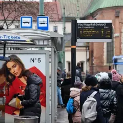 Ludzie czekają na przystanku Plac Wszystkich Świętych. Obok automatu z biletami miesięcznymi wisi tablica z rozkładem jazdy.