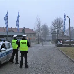 Policjanci stoją przy radiowozie przed bramą muzeum Auschwitz. Flagi Grecji powiewają na wietrze.