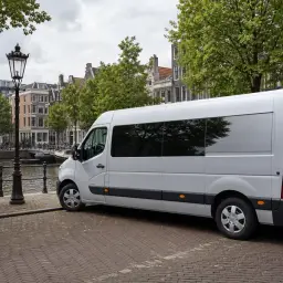Biały bus do Holandii zaparkowany przy kanale w Amsterdamie, obok rowerów i zabytkowych kamienic.