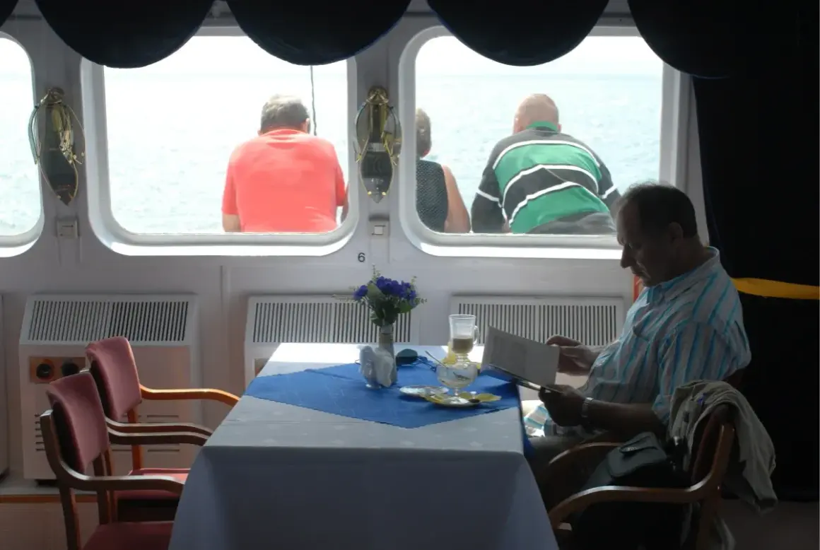 Rejs PKP Gdynia - Hel. Cena biletu? Mężczyzna czyta w restauracji na statku, podczas gdy inni pasażerowie patrzą na morze.