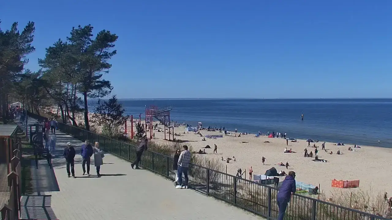 Ludzie spacerują promenadą w Stegnie, podziwiając plażę i morze. Słoneczny dzień zachęca do wypoczynku nad wodą.
