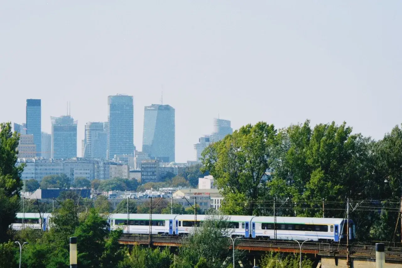 Pociąg PKP Intercity jedzie przez zielone drzewa, w tle widać warszawskie wieżowce. Zastanawiasz się, ile kosztuje bilet z Warszawy do Krakowa?
